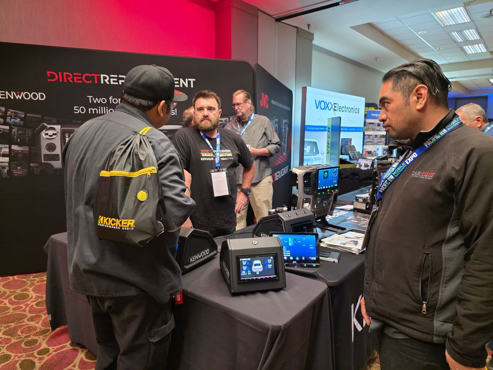 Three individuals gather around a booth at an exhibition, discussing technical equipment displayed on a table.