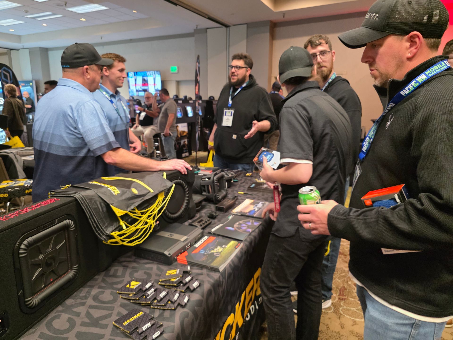 A group of people interact at a trade show booth featuring audio equipment, speakers, and promotional merchandise.