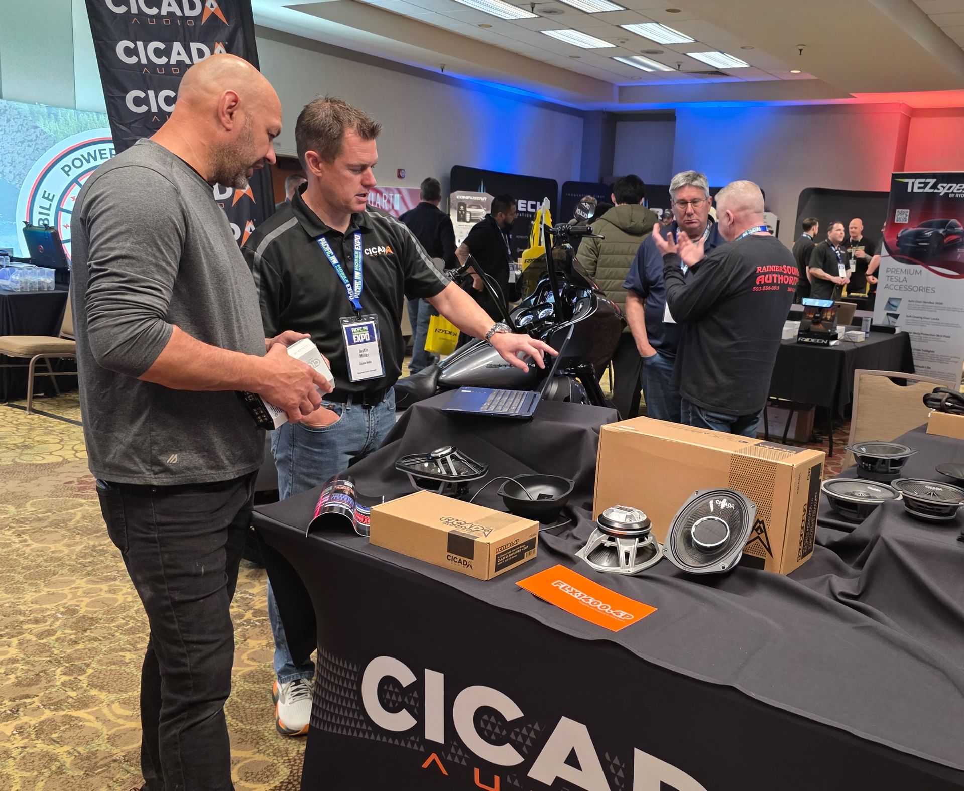 Two people discuss audio products at a Cicada Audio trade show booth in a convention hall.