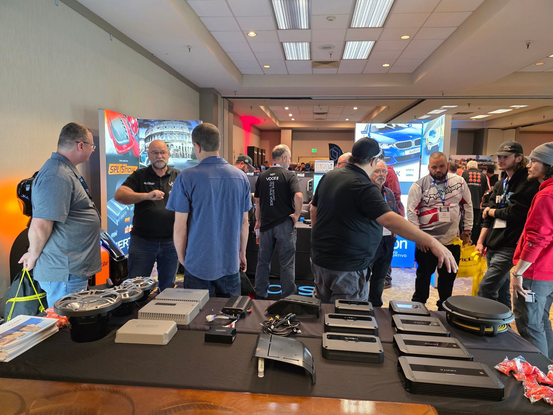 People gather around a table featuring car audio equipment and displays at a professional industry trade show event.
