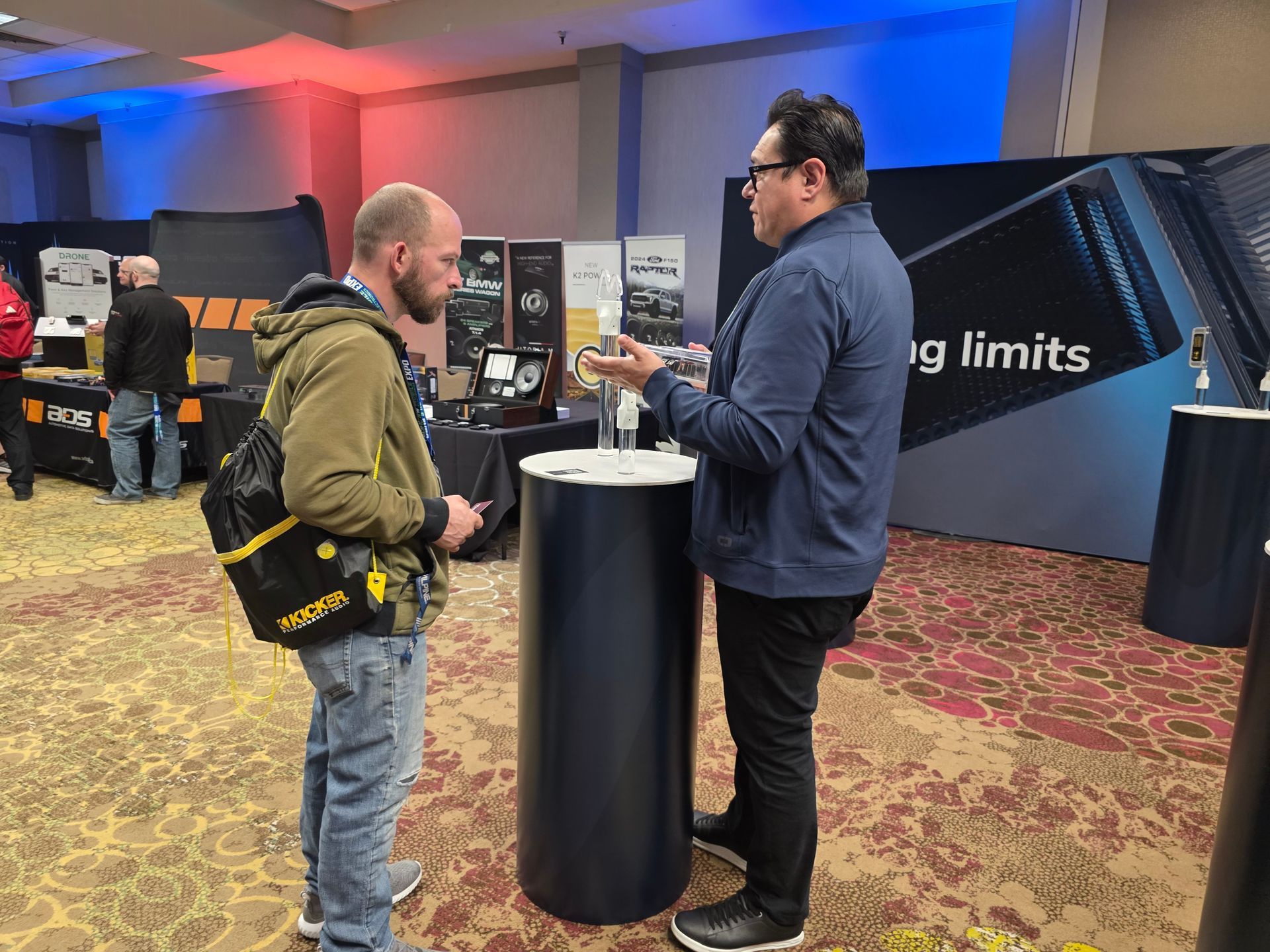Two people stand and talk by a circular pedestal at a professional trade show or conference.