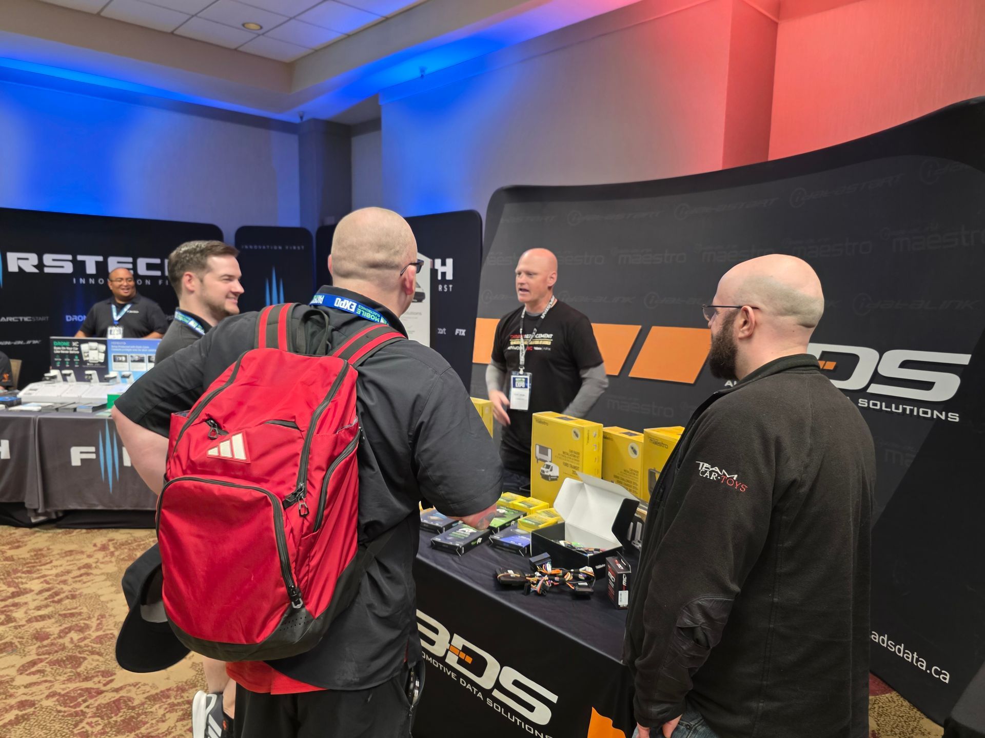 Four people at a trade show booth interacting, with a person in a red backpack facing away toward a branded black backdrop.