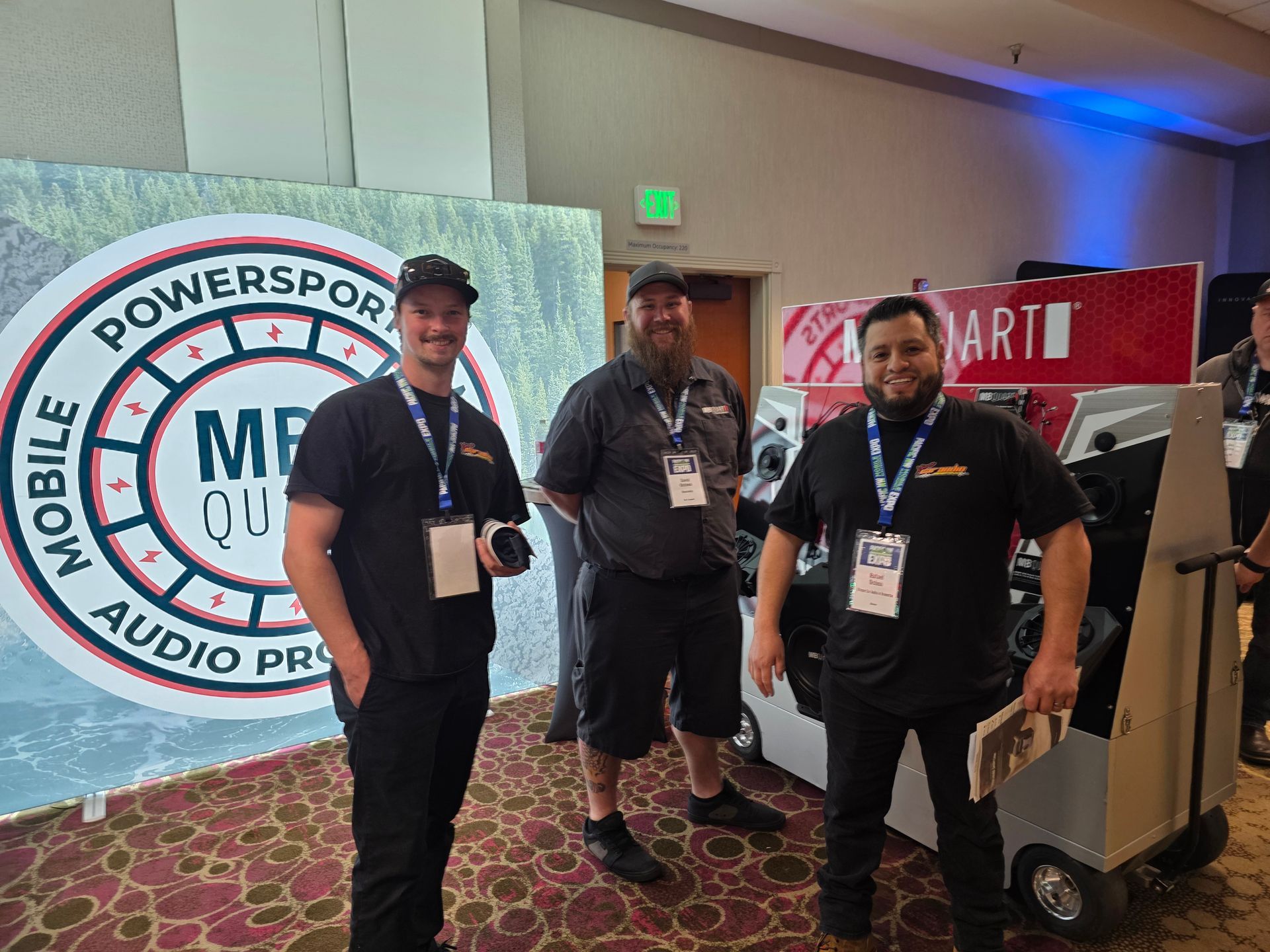 Three people stand in front of a Powersports mobile audio display at a convention, wearing black shirts and badges.