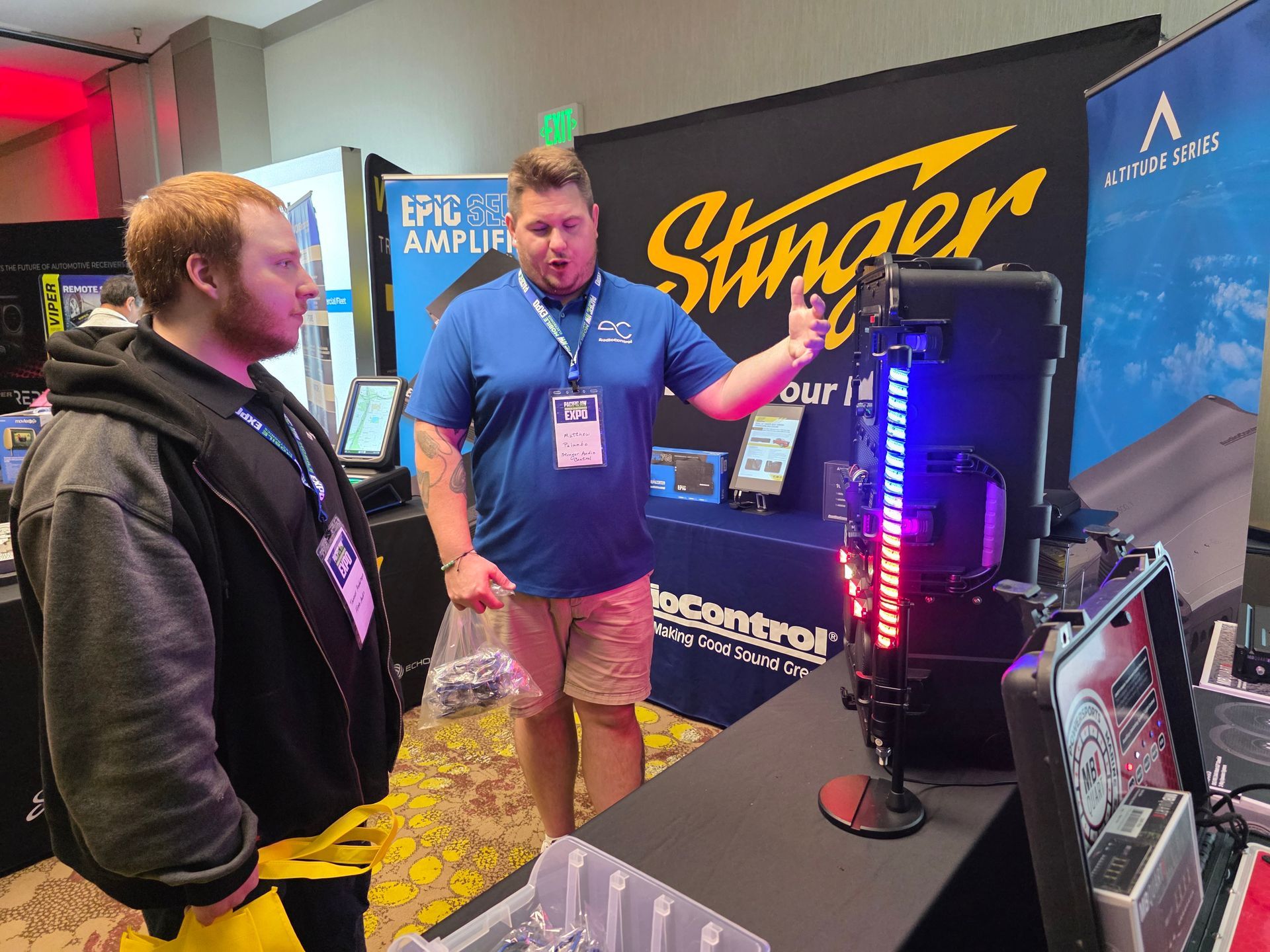 Two people at a booth discuss a display with vertical LED lights in front of a Stinger banner at an indoor event.