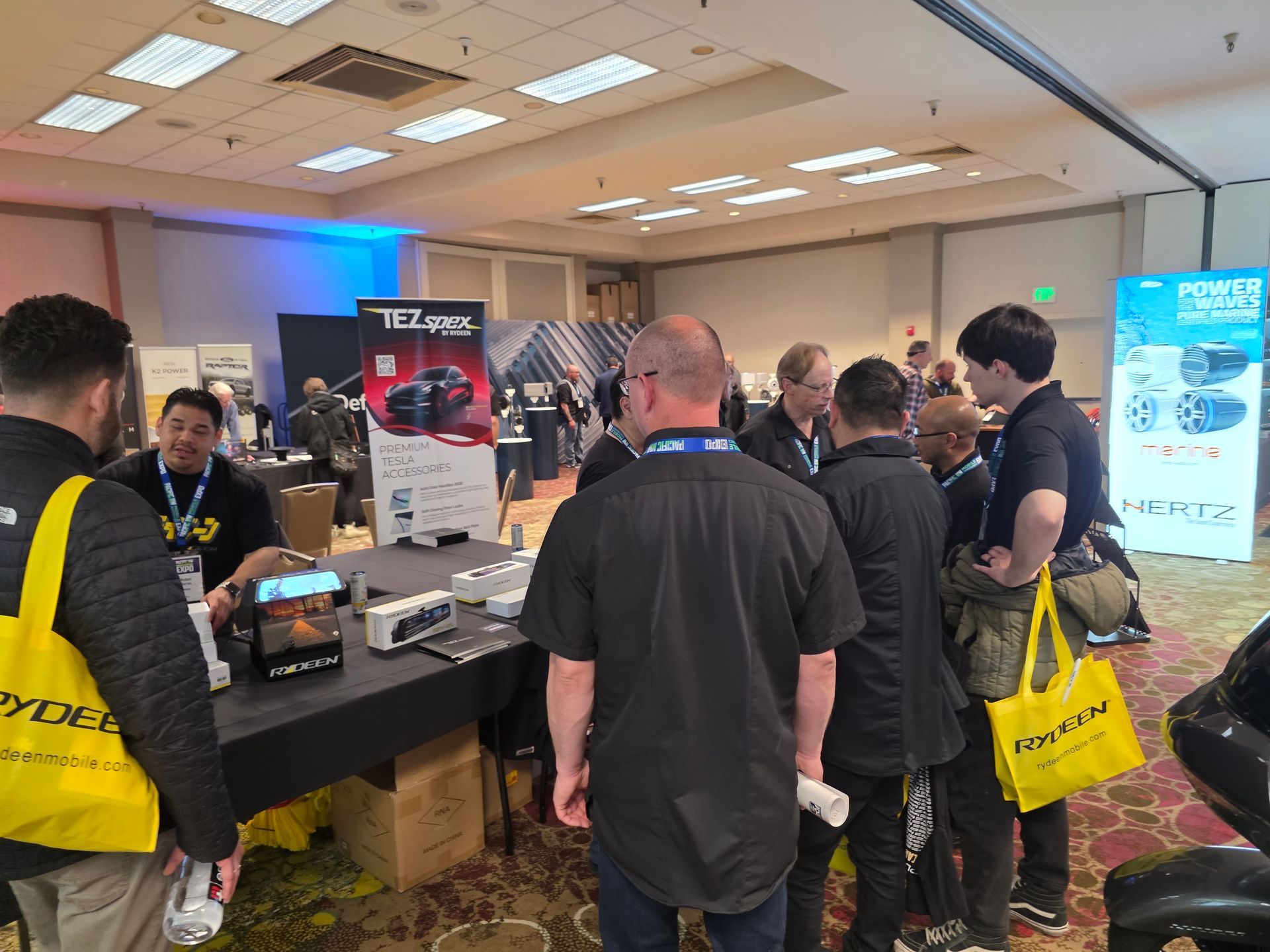 A group of people gathers around a trade show booth display featuring speakers, with a bright yellow bag in the foreground.