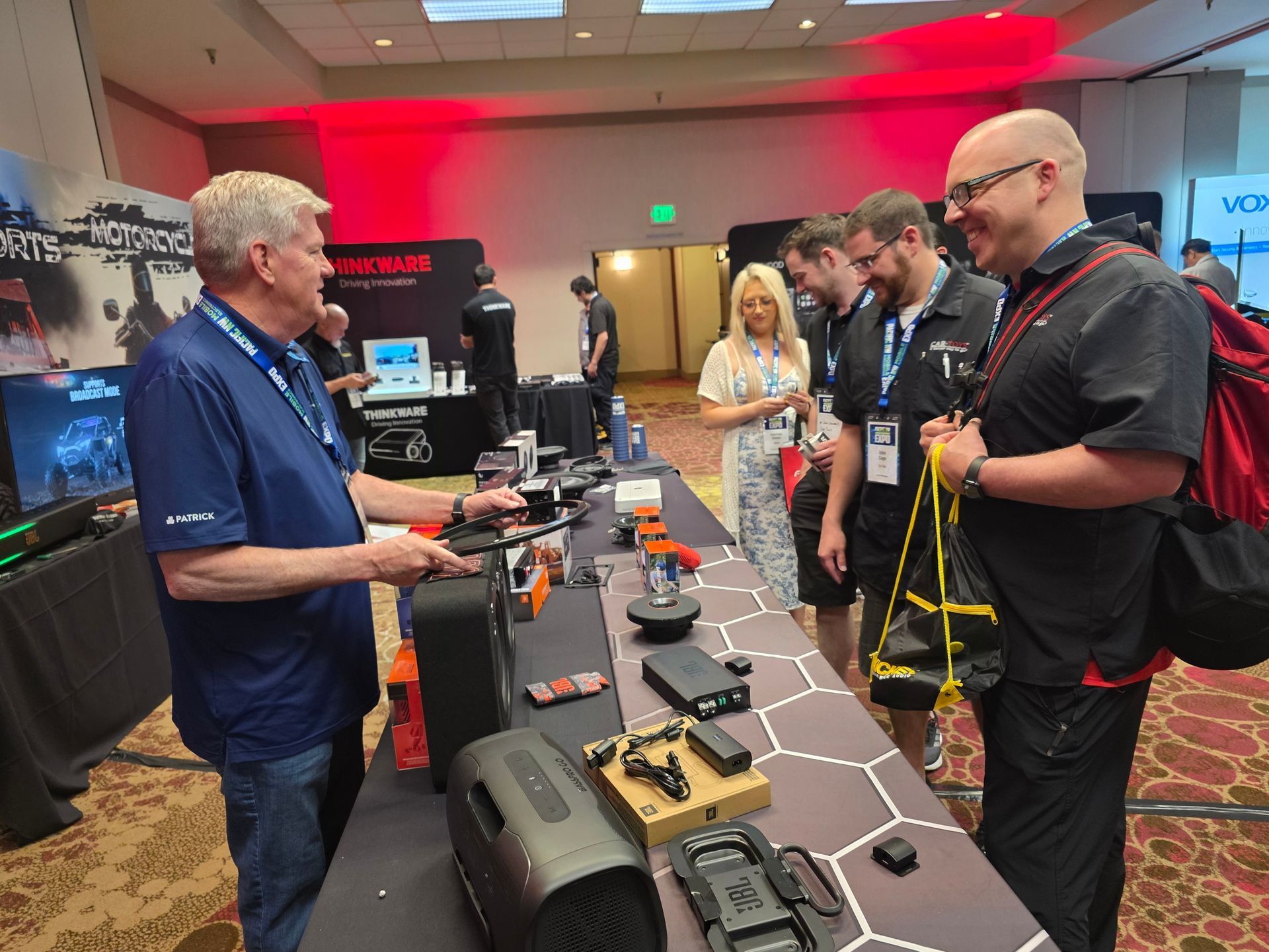 Exhibitor shows a portable speaker to attendees at a tech trade show booth with a patterned table and red lighting.