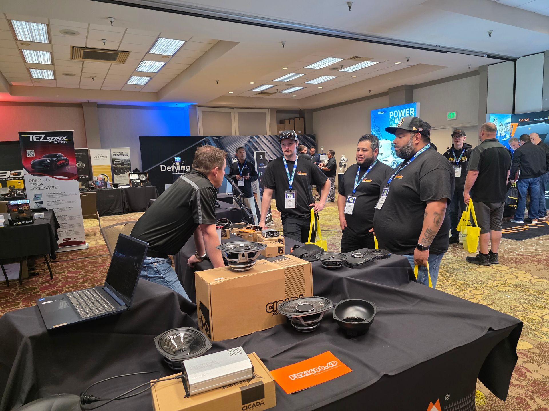 People stand around a trade show booth featuring audio equipment, including car speakers and a laptop on a table.