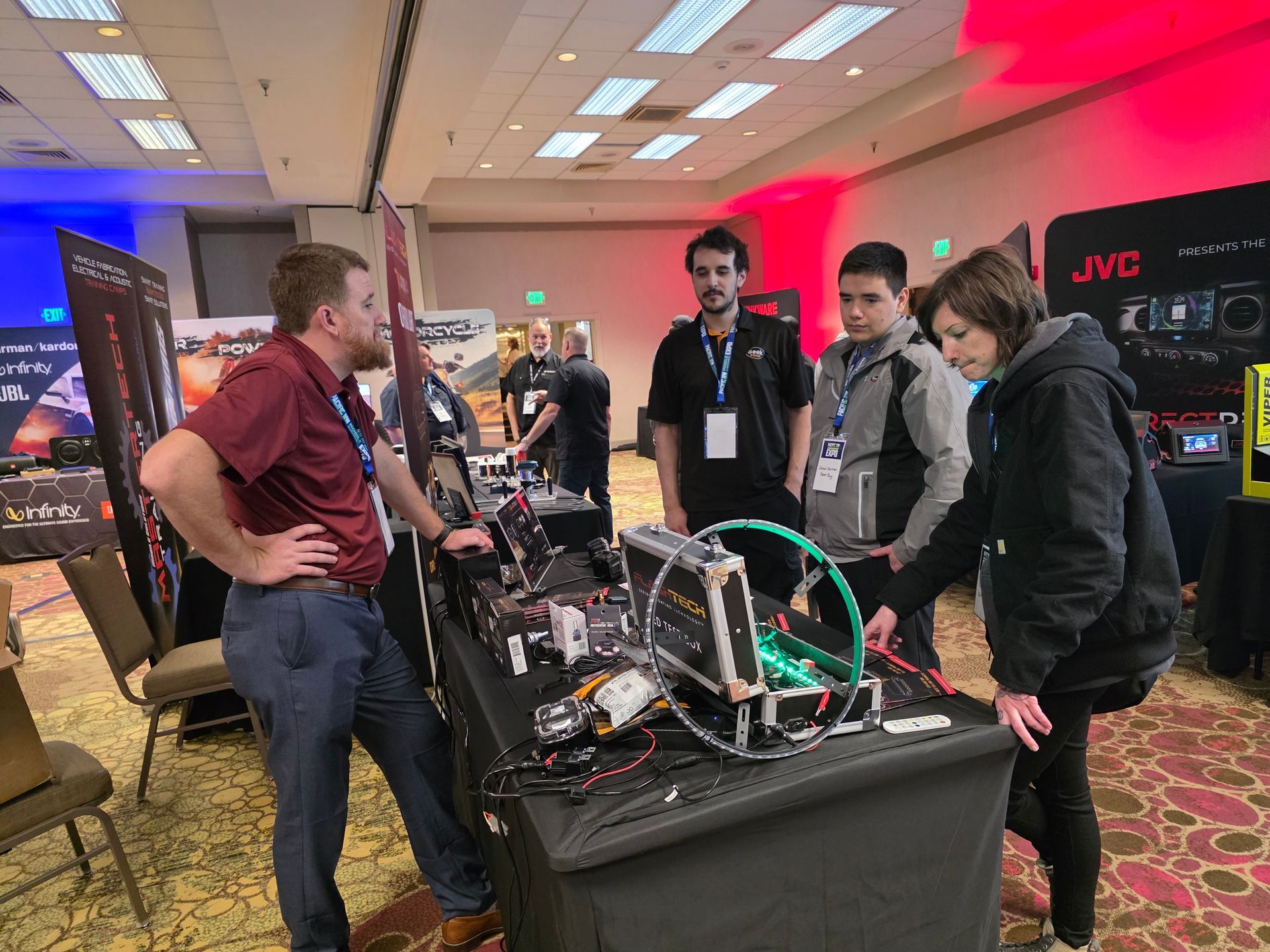 Four people gather around a demonstration table with electronic equipment in a large, brightly lit conference room.