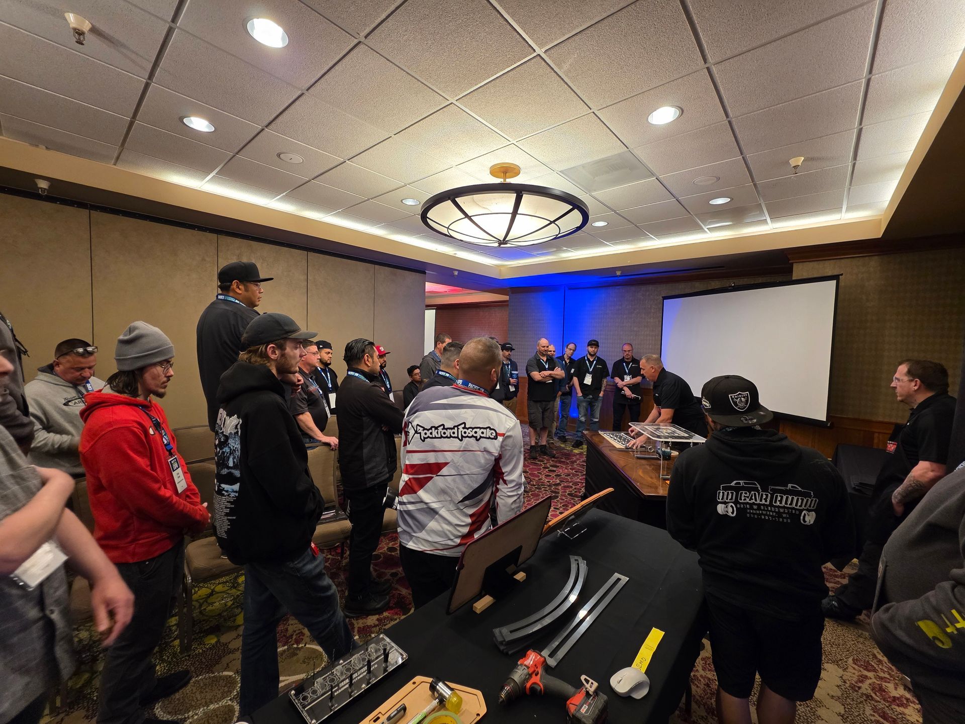 A group of people gathers in a meeting room, watching a demonstration at a table with tools and metal components.
