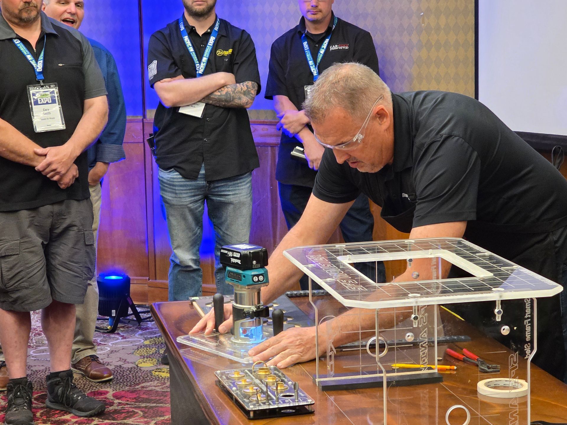A person demonstrates the use of a handheld power router on a clear acrylic jig at a table, while others observe.