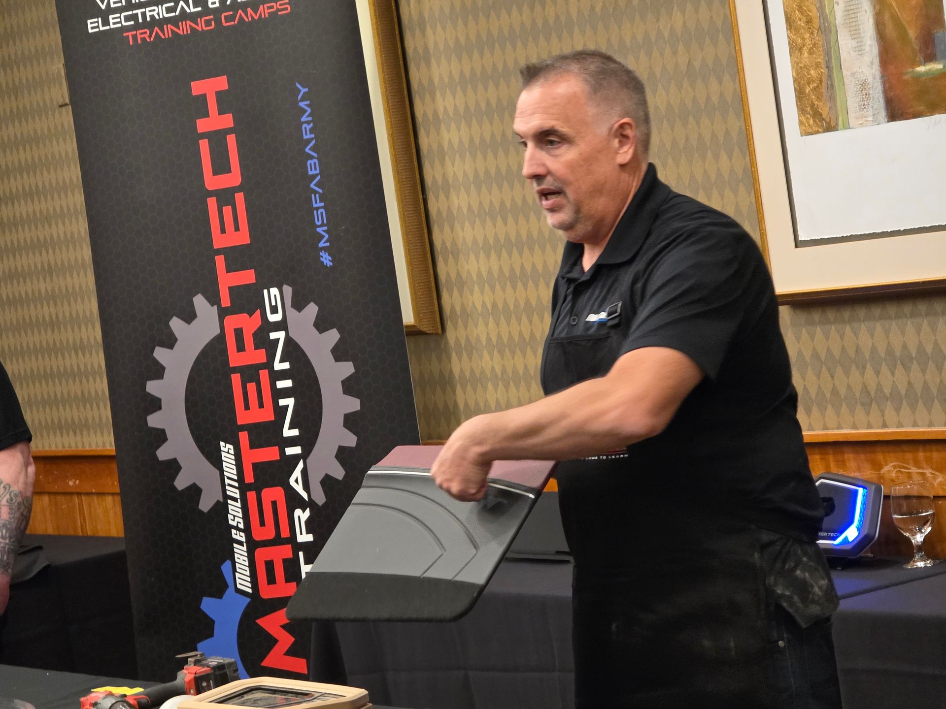 A person in a black polo shirt holds a grey training panel in front of a Mastertech Training banner in a conference room.