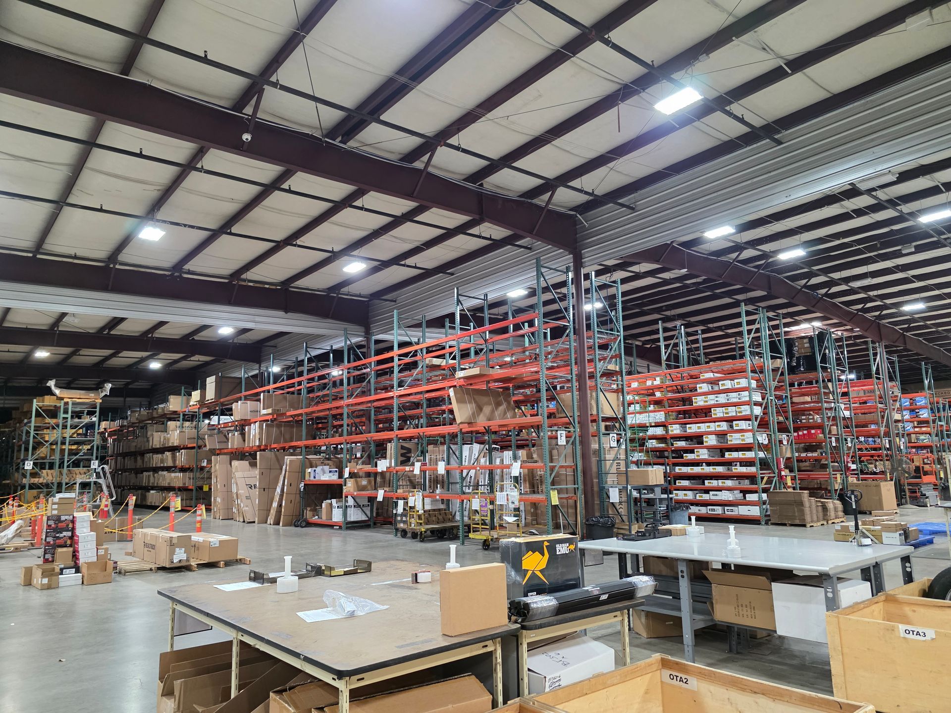 A large warehouse featuring rows of high metal shelving stocked with boxes and goods, with work tables in the foreground.