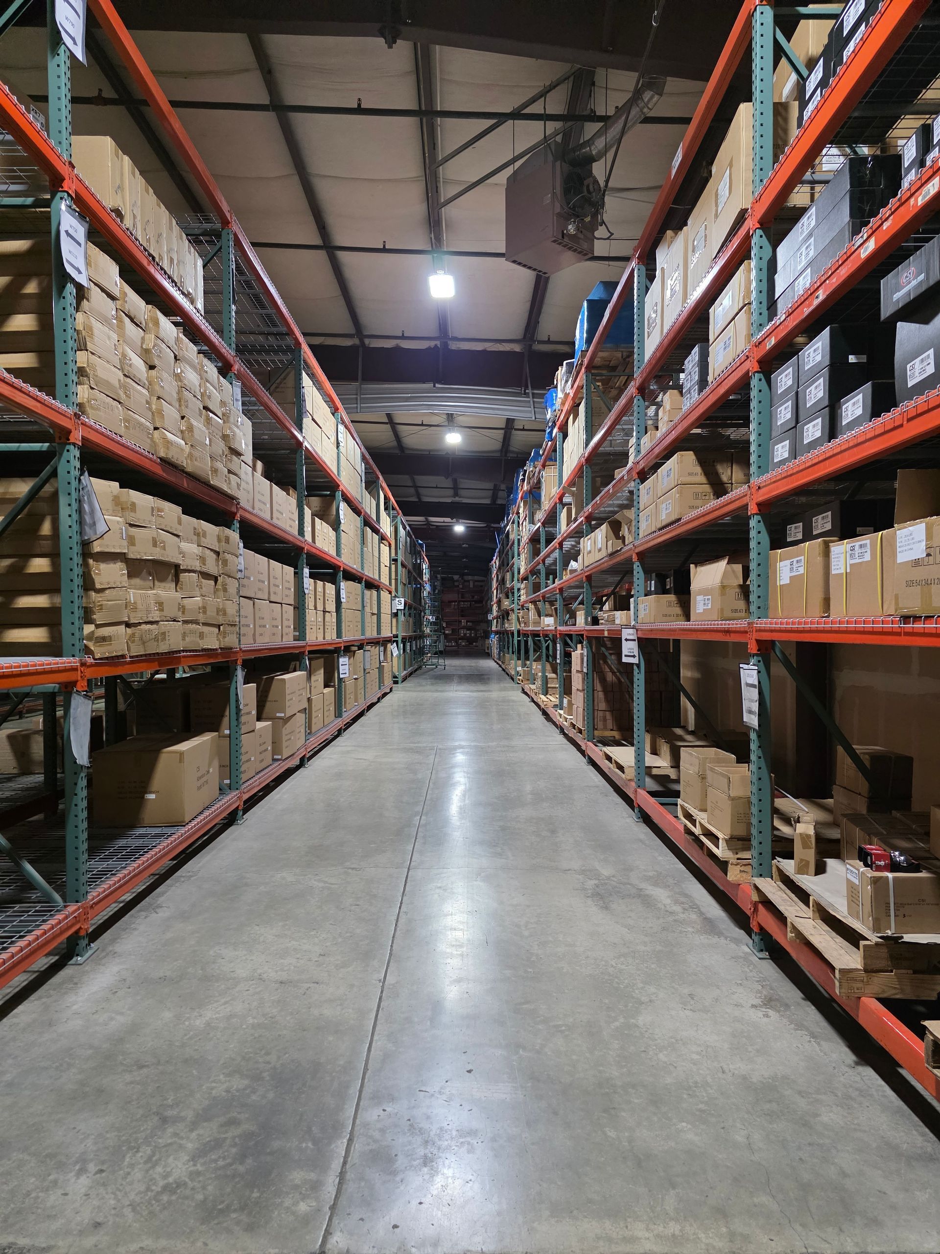 A warehouse aisle with tall, orange metal shelving units filled with cardboard boxes on either side.