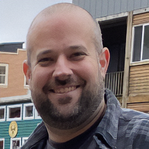 A bearded man smiles while sitting in a classroom or conference setting with other people in the background.
