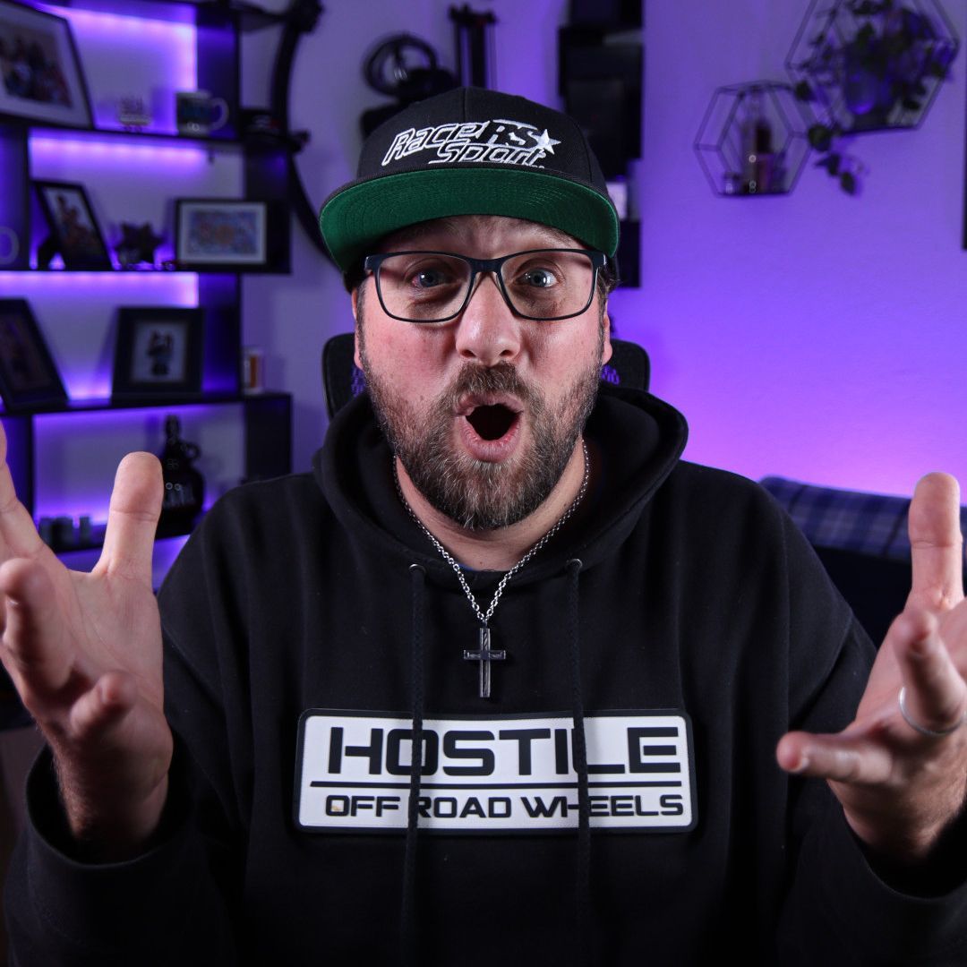 A bearded man in a Hostile Off-Road Wheels hoodie and cap with glasses looks surprised with hands raised in a lit studio.