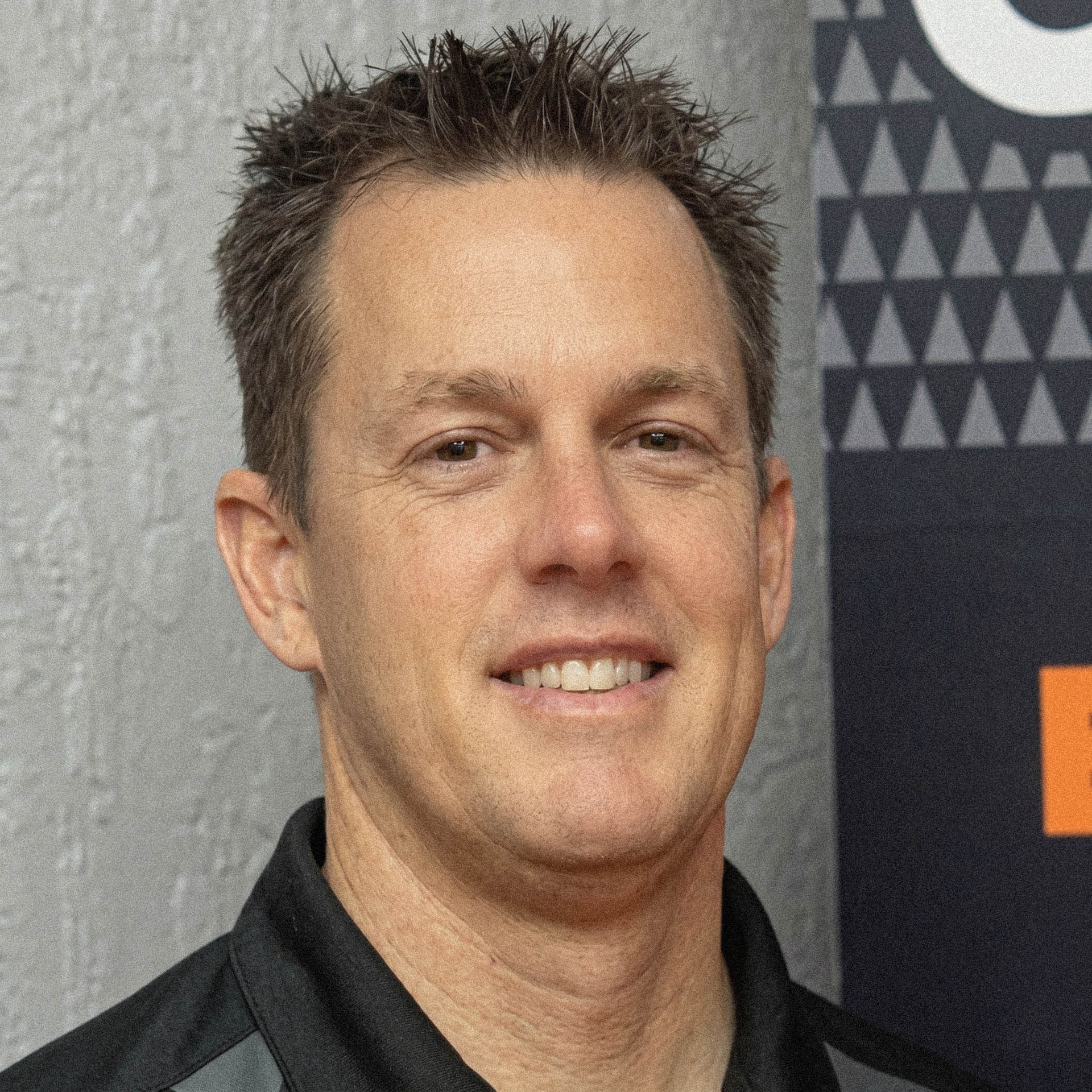 Man smiling in a dark shirt against a gray and black patterned backdrop