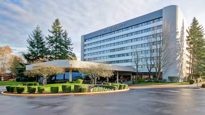 A tall, grey multi-story hotel building with a curved wing, a front drive, and landscaped greenery on a sunny day.
