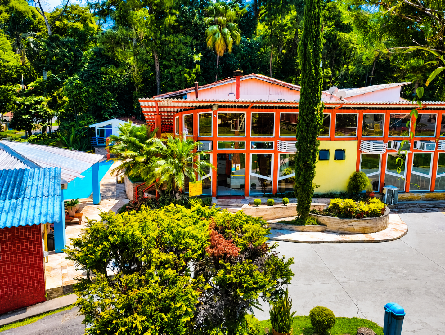 Edifícios tropicais coloridos em torno de uma entrada de automóveis curva, com árvores e vegetação exuberante ao fundo.