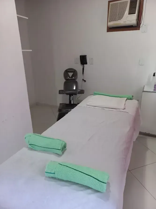 Sala de tratamento com uma maca branca, toalhas verdes e uma cadeira preta ao lado de um ar-condicionado fixado na parede.