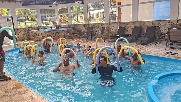 Crianças usando macarrões de piscina amarelos em uma piscina coberta durante uma aula.