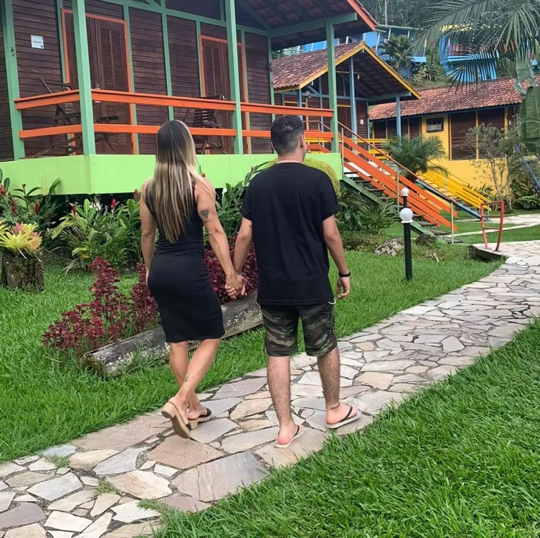 Casal de mãos dadas em um caminho de pedra ao lado de cabanas coloridas de um resort e vegetação exuberante.