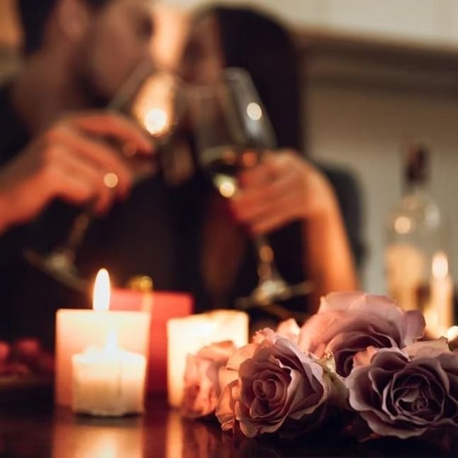 Velas e rosas sobre uma mesa de jantar com duas pessoas brindando com vinho ao fundo.