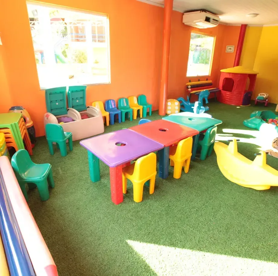 Sala de jogos iluminada com mesas, cadeiras e brinquedos coloridos para crianças, além de um tapete verde sob paredes laranja.