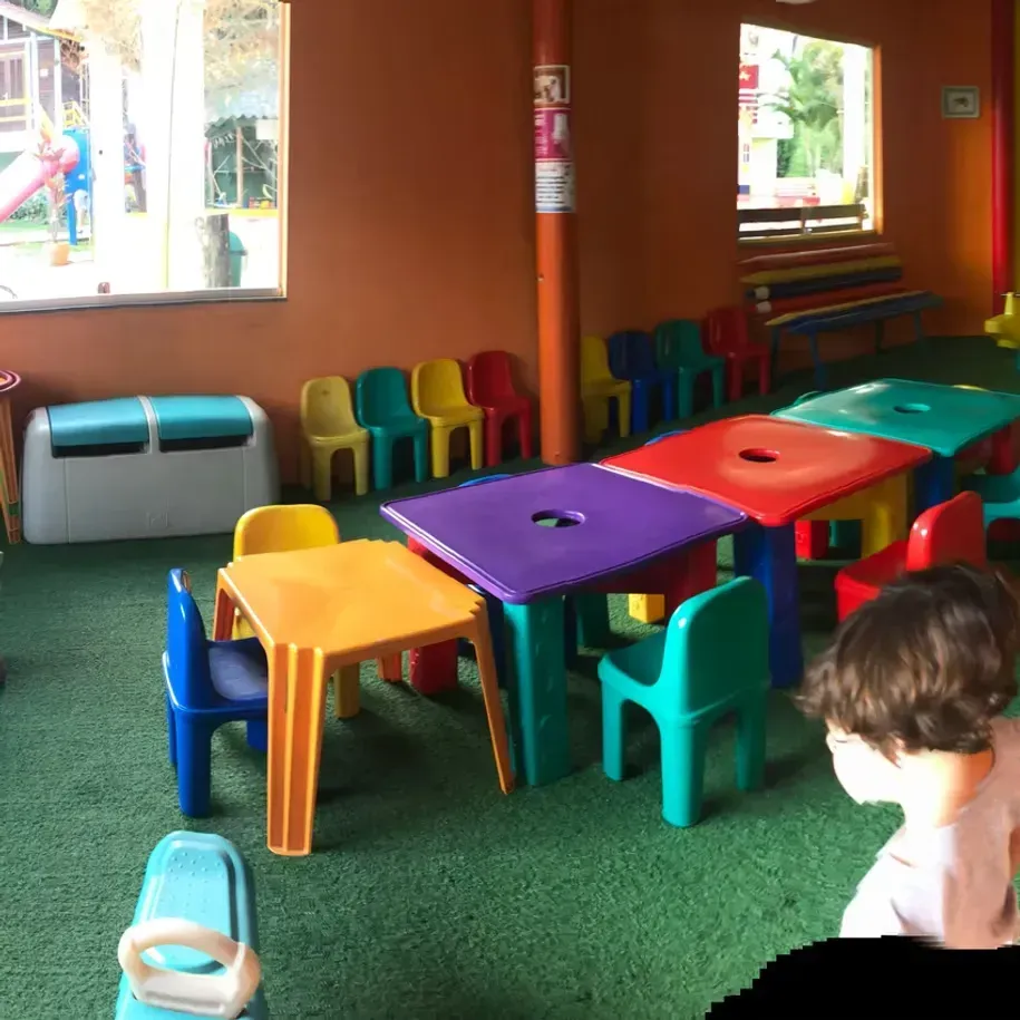 Mesas e cadeiras coloridas em tamanho infantil em uma sala de jogos, com uma criança sentada na extremidade direita.