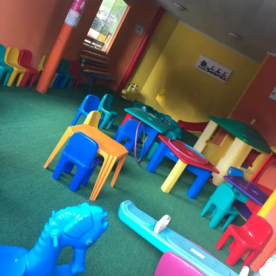 Sala de jogos infantil colorida com mesinhas e cadeiras sobre um tapete verde, vista inclinada.