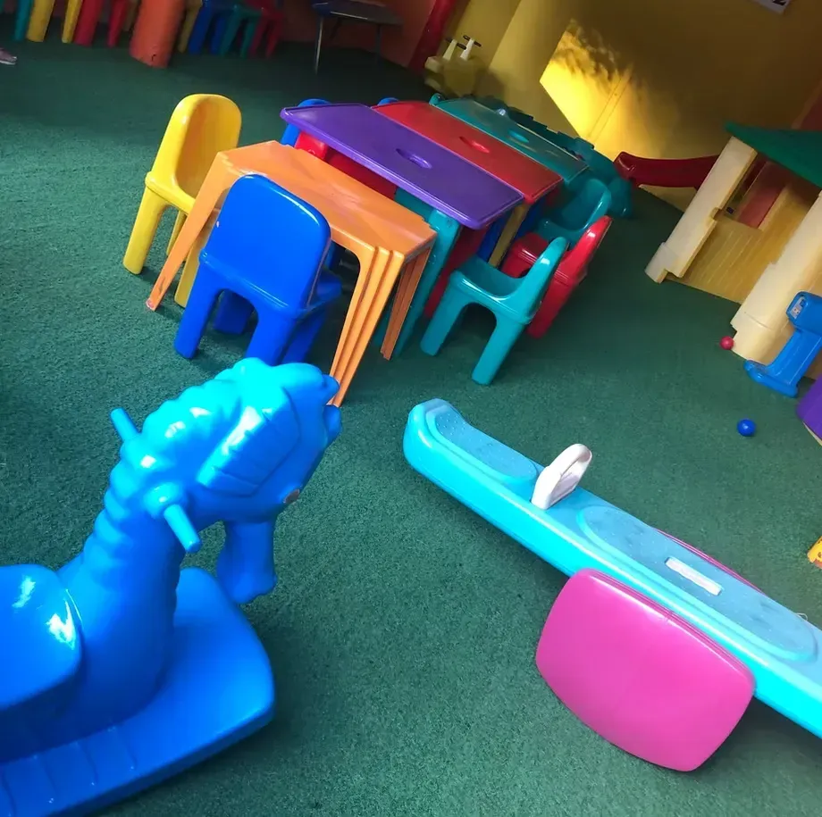 Cadeiras e mesas infantis de plástico coloridas em uma sala de jogos com carpete verde.