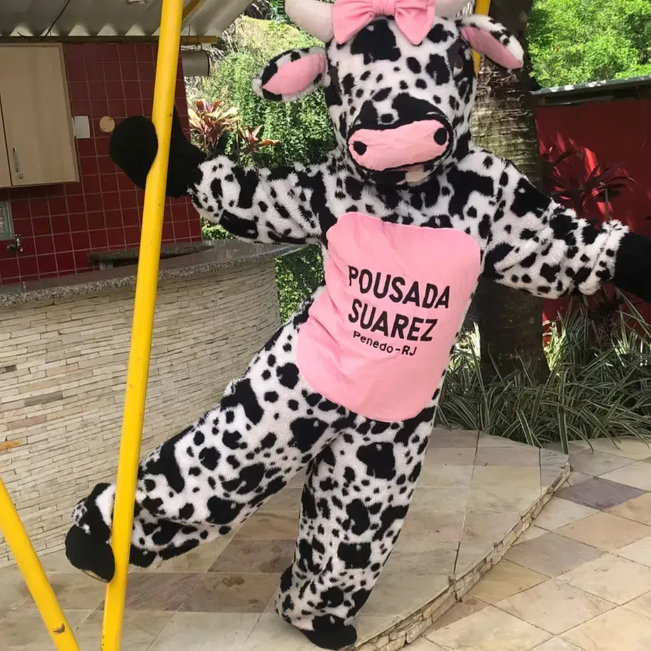 Mascote dálmata de avental rosa em pé dentro de uma pousada, segurando uma vara amarela na entrada.
