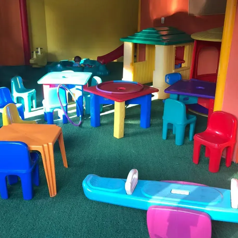 Área de recreação interna colorida com cadeirinhas de plástico, mesas e escorregadores de brinquedo sobre carpete verde.