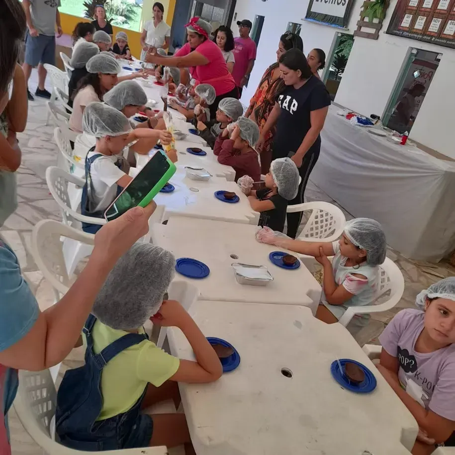 Pessoas com toucas de cabelo sentadas em uma longa mesa com pratos, comendo ou servindo comida em um refeitório iluminado.