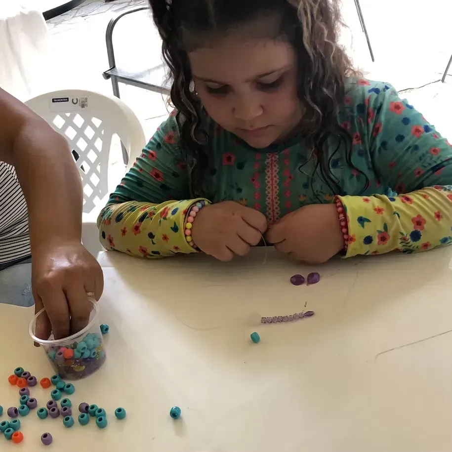 Criança fazendo artesanato colorido com miçangas em uma mesa, concentrada em colocar pequenas miçangas com as mãos.