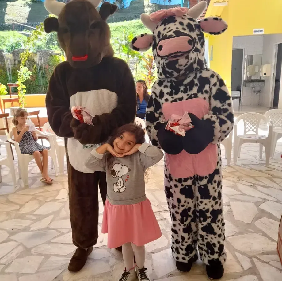 Criança posando com duas pessoas vestidas de vaca em um ambiente interno, segurando presentes cor-de-rosa, com cadeiras e um pátio iluminado ao fundo.