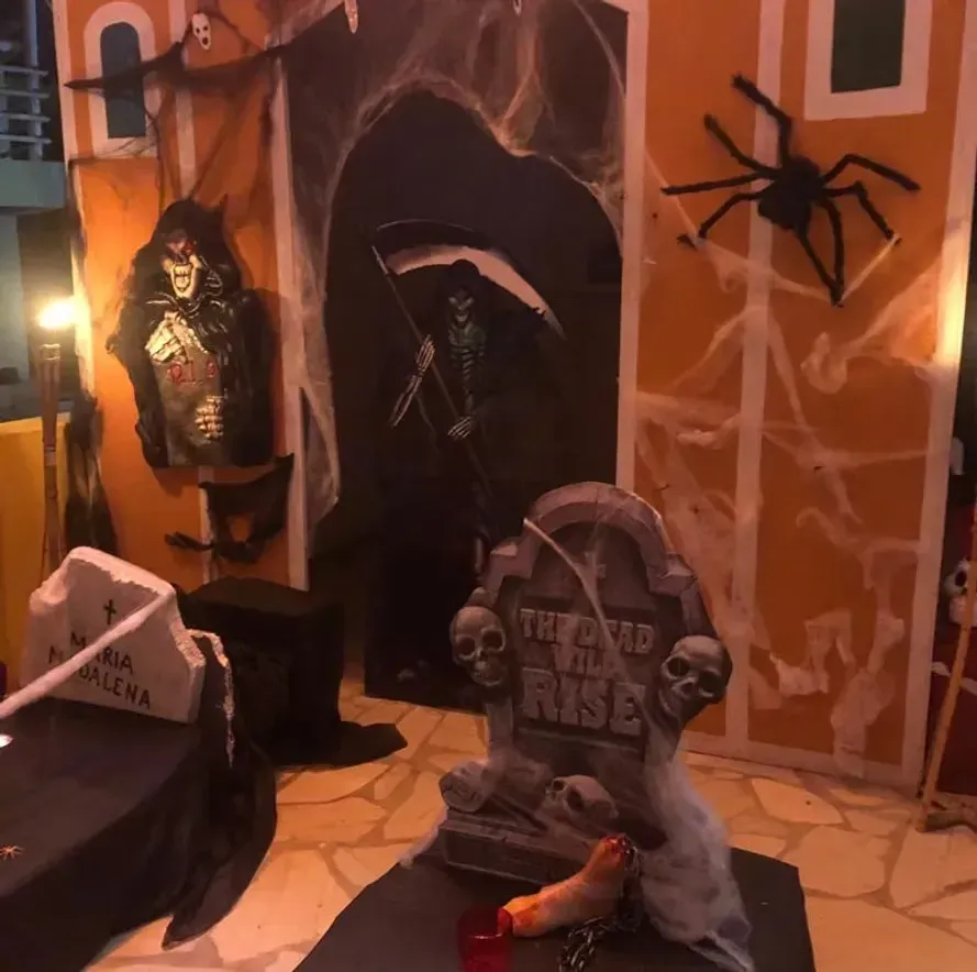 Decoração de cemitério para o Halloween com esqueletos brilhantes, lápides e uma aranha gigante em uma porta iluminada de laranja.