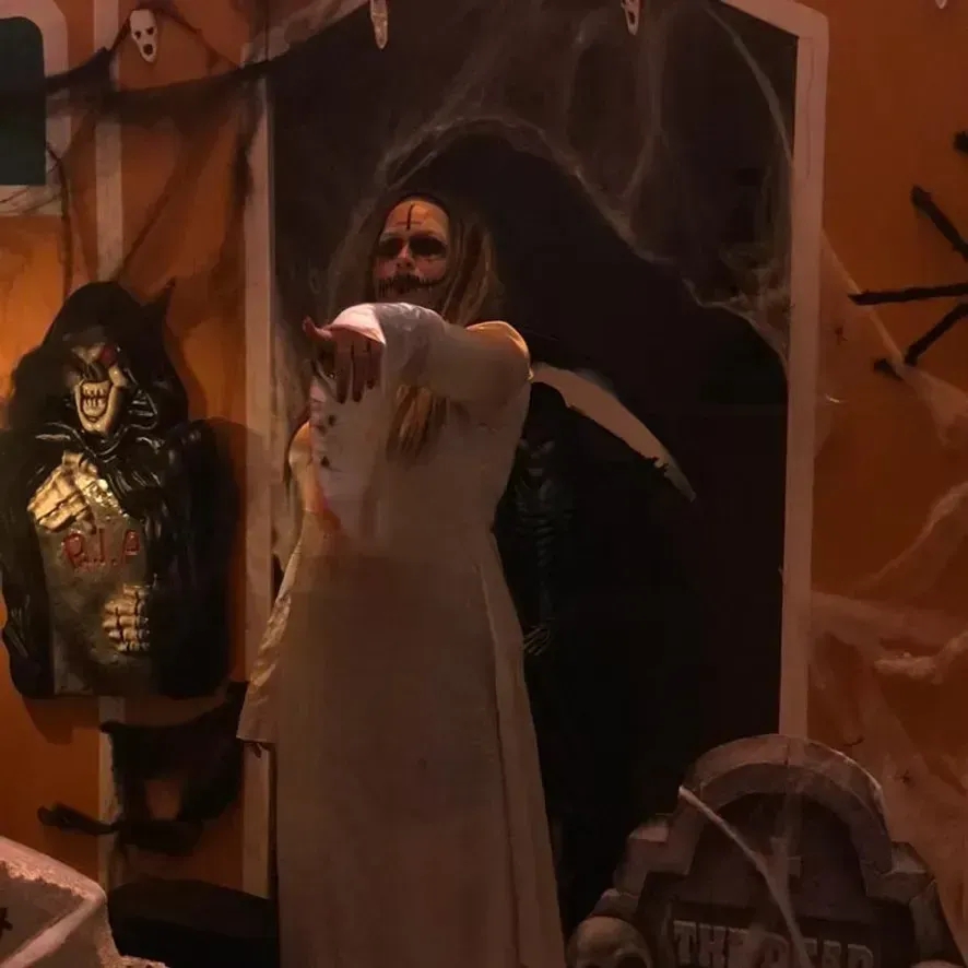 Cena de Halloween com uma figura fantasmagórica apontando para a câmera, emoldurada por teias de aranha e decoração de parede assustadora.