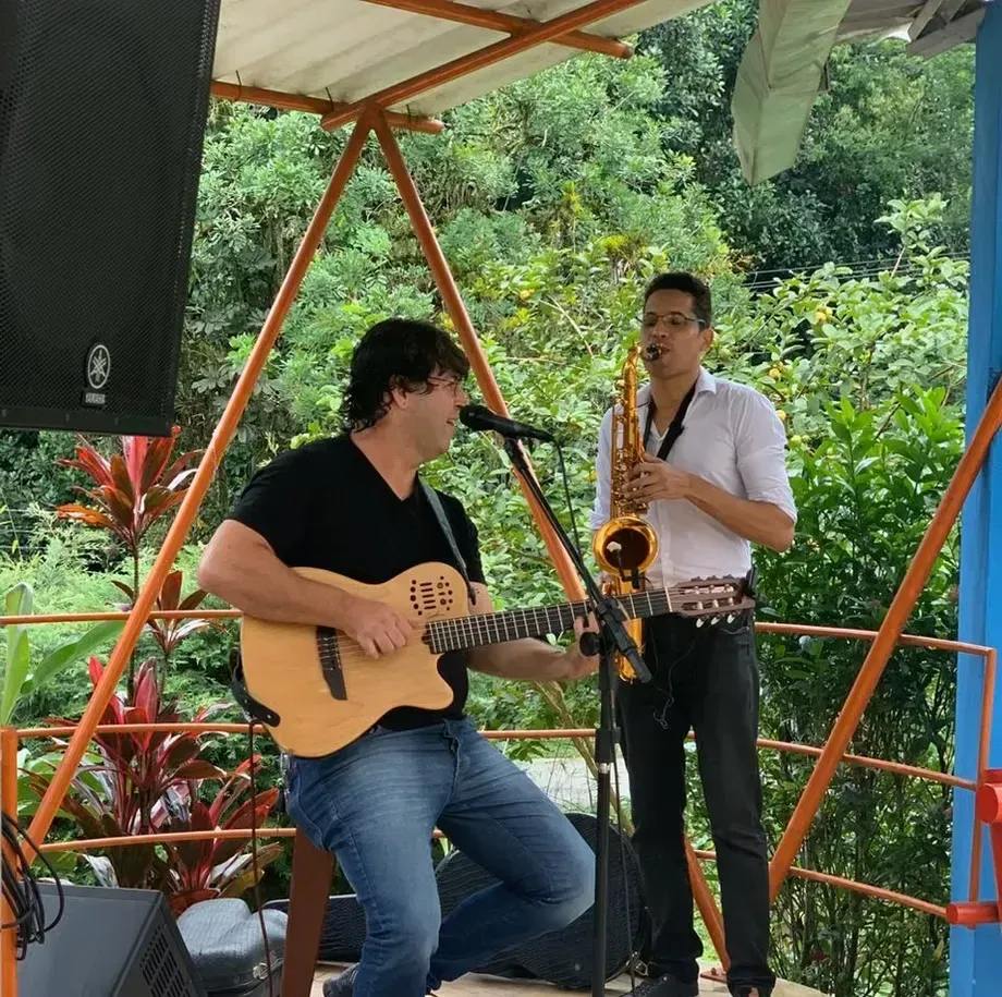 Dois músicos se apresentam em um deck externo coberto, um tocando guitarra e o outro saxofone.