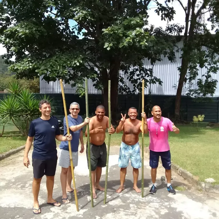Cinco pessoas em pé ao ar livre, num gramado, segurando longas varas de madeira e sorrindo perto de uma grande árvore.