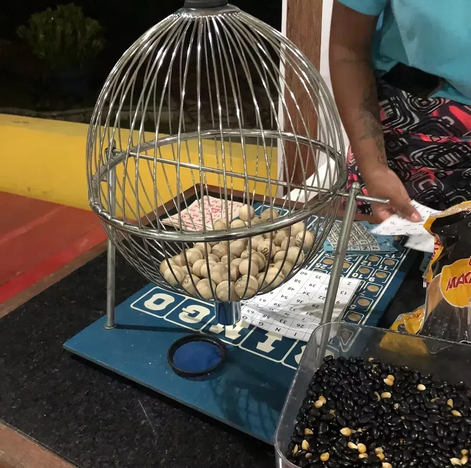 Tambor de loteria com gaiola de arame e bolas numeradas sobre uma mesa ao lado do braço de uma pessoa e uma bandeja com fichas pretas e douradas.