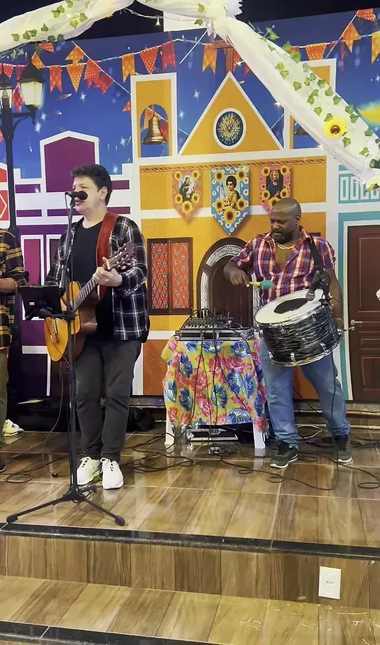 Músicos se apresentando em um palco colorido, um tocando guitarra e cantando, outro tocando bateria.