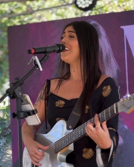 Músico tocando guitarra elétrica branca no palco, com uma cantora desfocada ao fundo.