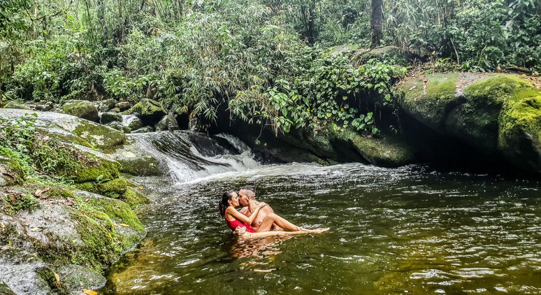 Pessoa de maiô vermelho relaxando em um riacho rochoso na floresta, cercada por folhagem verde.