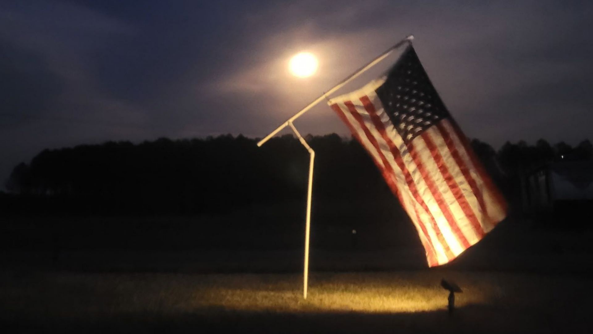 American flag lit by a spotlight at night, waving on a pole in a dark field