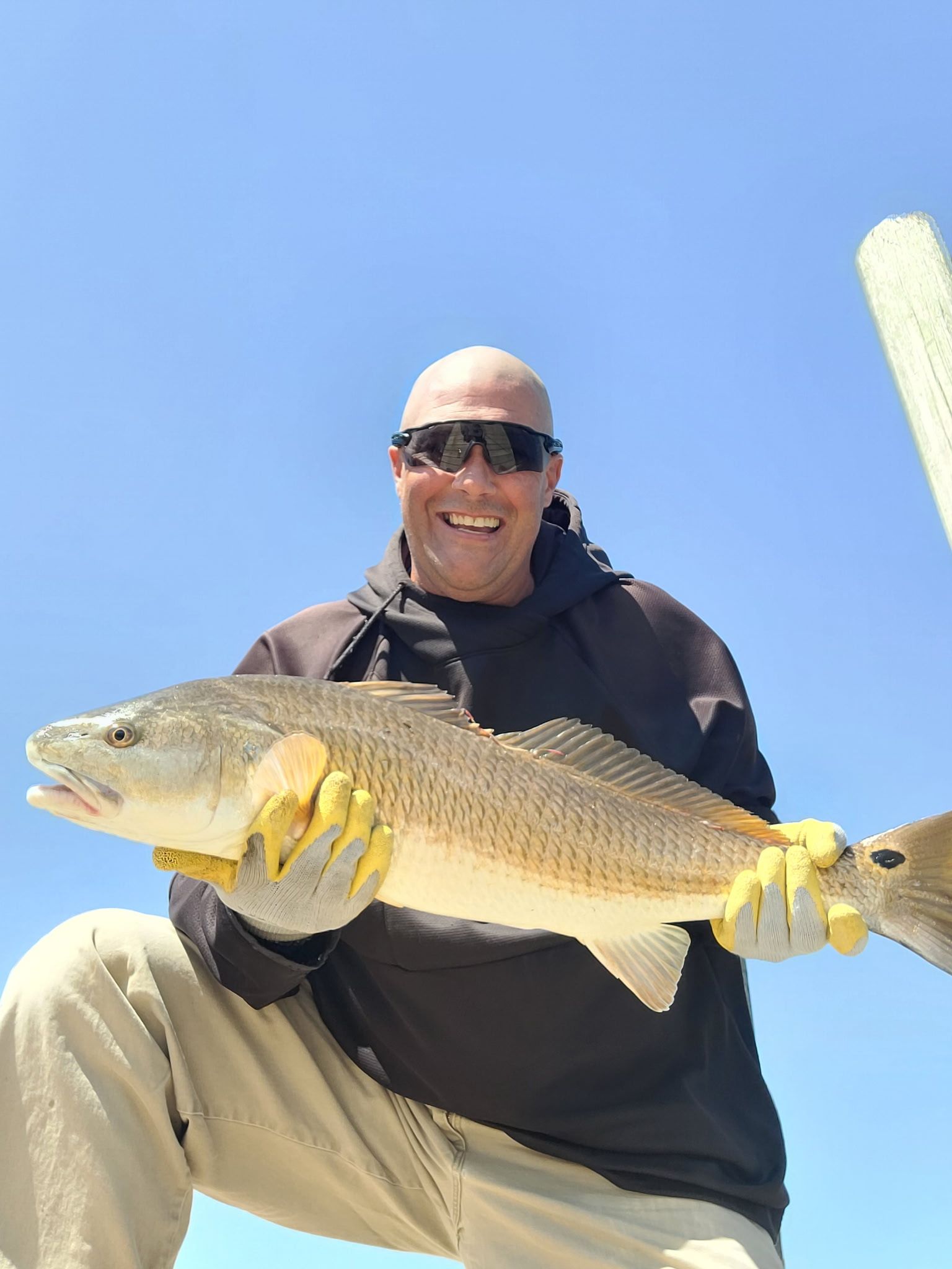 Man holding a large fish outdoors under a clear blue sky, smiling in sunglasses and gloves