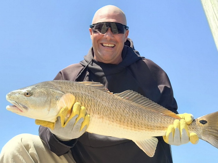Person holding a large fish against a blue sky, smiling and wearing sunglasses and gloves