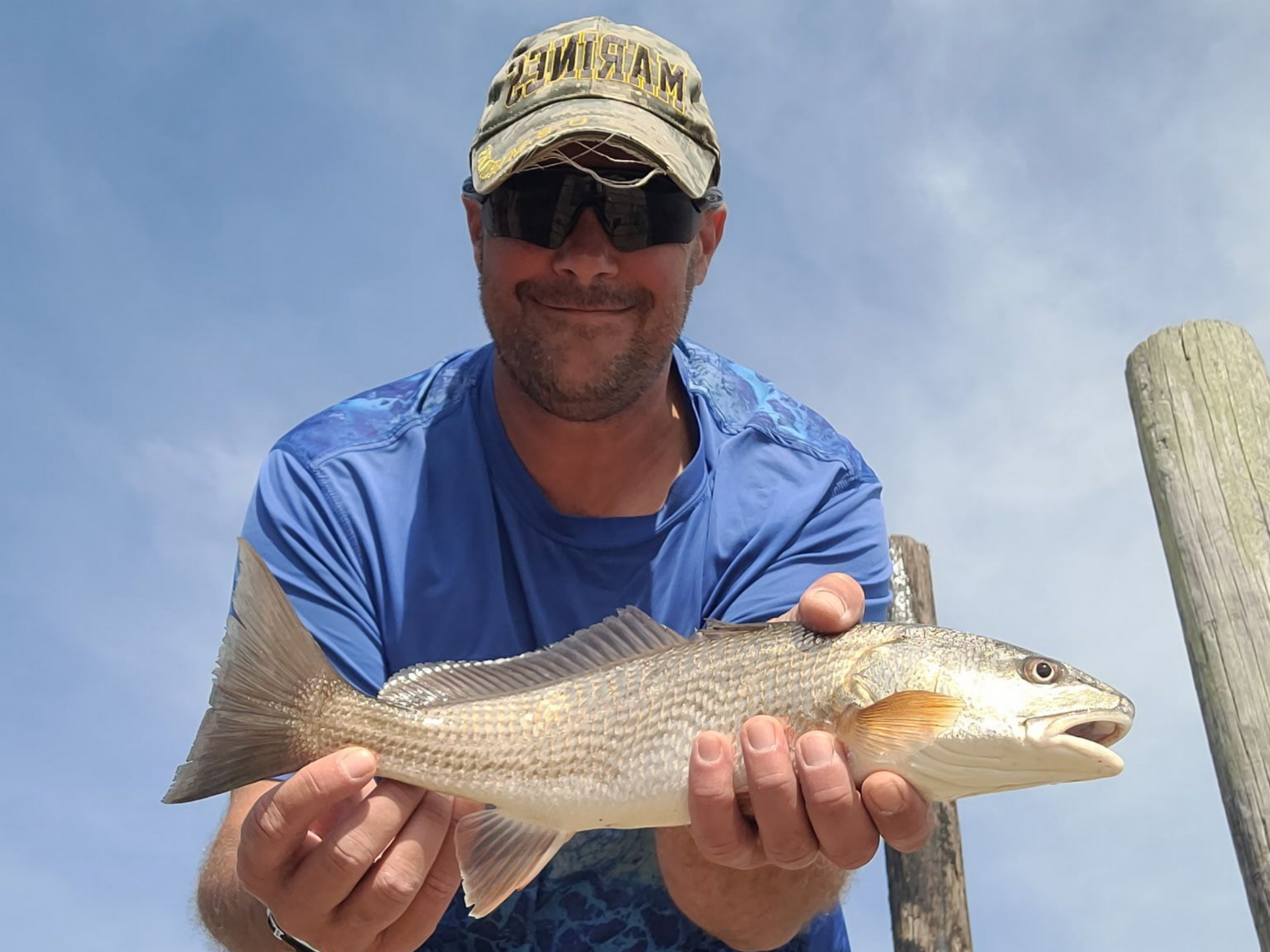 Man holding a silver fish outdoors against a blue sky, wearing a cap and sunglasses.