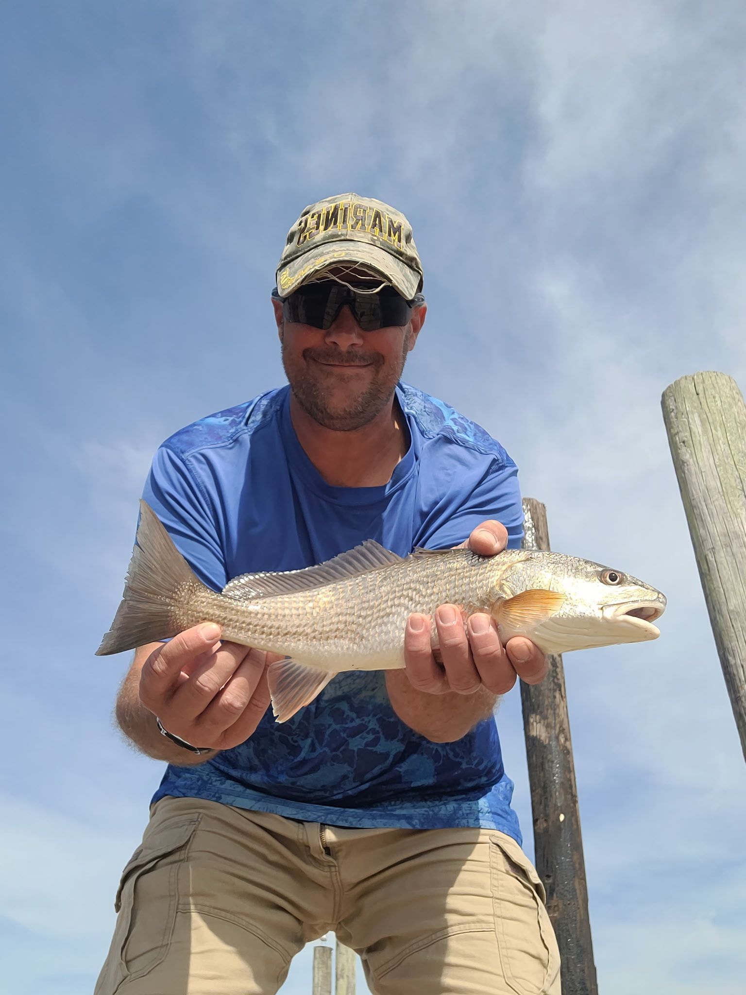 Man holding a fish outdoors against a blue sky, wearing a blue shirt and cap