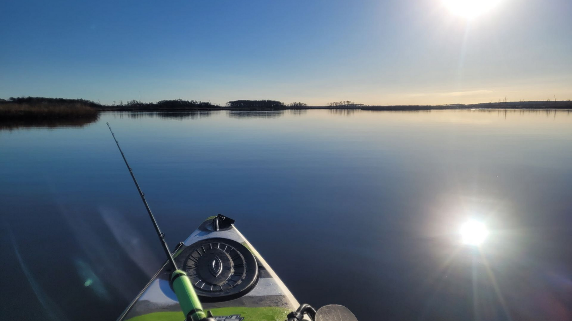 Kayak bow with fishing rod on a calm lake at sunrise, with bright sun reflections and distant islands