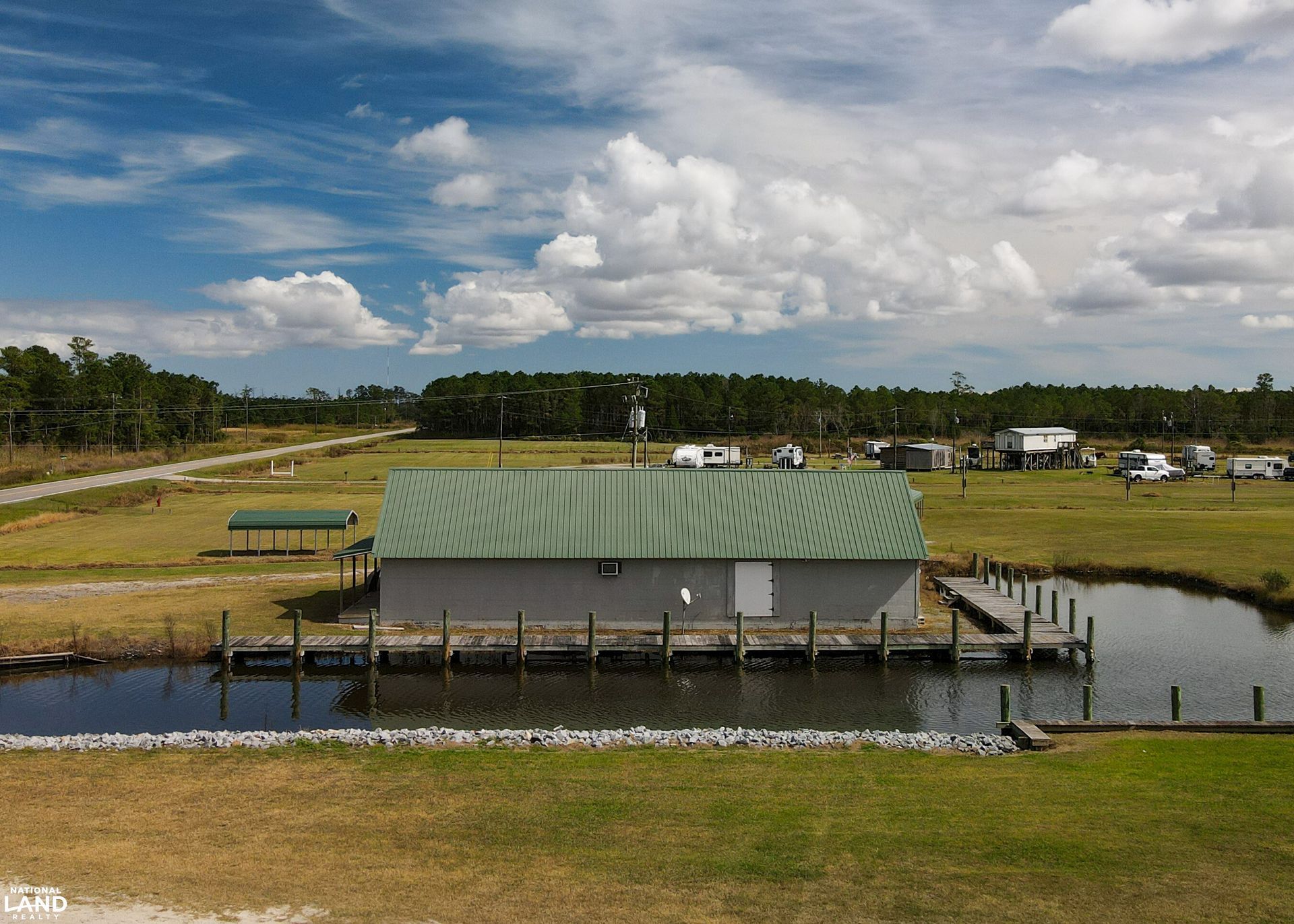 Rural pond with a green-roofed dock building under a cloudy sky, surrounded by grassy fields.